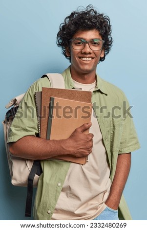 Vertical shot of cheerful curly haired Hindu teenage guy smiles gladfully poses with notepads carrying backpack ready for studying comes on English class for tutor poses against blue background. Royalty-Free Stock Photo #2332480269