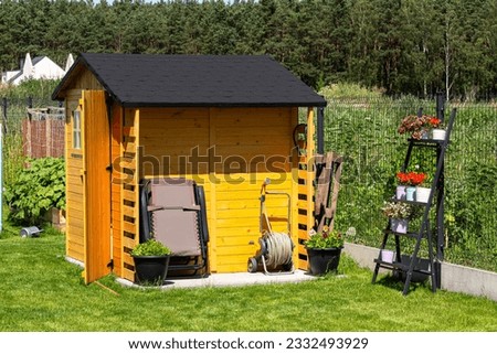 A wooden garden shed standing on a concrete foundation in a garden, flowers and tools visible. Royalty-Free Stock Photo #2332493929