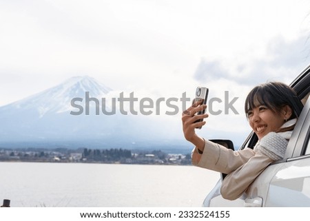 Happy Asian woman enjoy and fun outdoor activity lifestyle using mobile phone taking selfie with Mt Fuji covered in snow during travel nature road trip on car at Kawaguchi lake on holiday vacation.