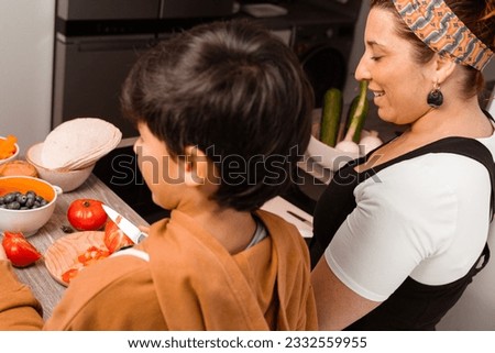 A mom cooks at home with her brown-haired, 10-year-old son who is chopping a tomato-they are in a sunny kitchen-Pictures of family members cooking-Family cooking