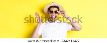 Concept of tourism and holidays. Close-up of surprised man shouting for joy, enjoying vacation, wearing sunglasses with summer hat, yellow background.