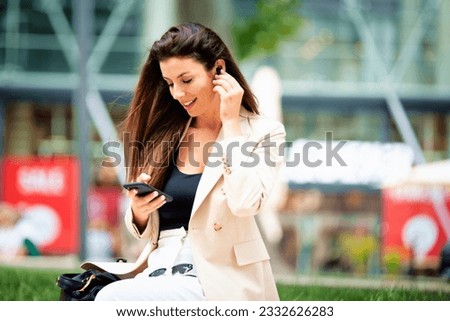 Attractive brunette haired woman with earphones is sitting on a bench in the street and using phone. Smiling female wearing business casual and sunglasses and having video call.  Royalty-Free Stock Photo #2332626283