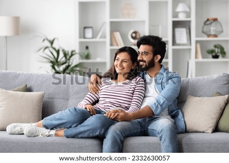 Loving cheerful eastern millennial man and woman in comfy casual outfit cuddling at home, happy sweet couple sitting on couch in living-room, embracing and looking at copy space. Domestic lifestyle Royalty-Free Stock Photo #2332630057