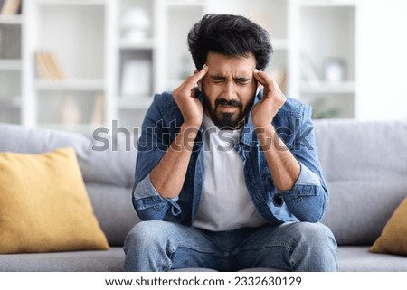 Portrait of young indian guy suffering from migraine at home, eastern man feeling unwell, touching his temples with closed eyes, suffering acute headache while sitting on couch, copy space Royalty-Free Stock Photo #2332630129
