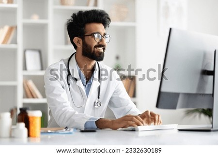 Focused young indian doctor in white medical uniform and eyeglasses sit at desk in hospital, work on laptop online. Arab man general practitioner look at computer screen, consult patient Royalty-Free Stock Photo #2332630189