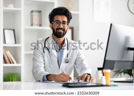 Friendly smiling eastern handsome young man wearing medical coat, eyeglasses with stethoscope on his neck family doctor working at his office at modern clinic, writing prescription, copy space