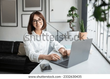 Successful freelancer. Happy young lady sitting at desk with laptop, cheerful european woman smiling and posing at camera while working on computer in cozy office, free space Royalty-Free Stock Photo #2332630435