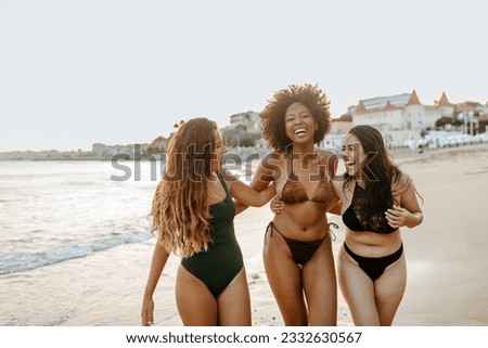 Excited diverse female friends in swimwear embracing and laughing cheerfully while walking at the beach, enjoying their free time on ocean shore