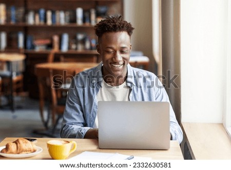 Happy cheerful handsome young black guy in casual outfit working online on laptop computer at cozy city cafe, entrepreneur responding emails while have breakfast at restaurant, copy space Royalty-Free Stock Photo #2332630581