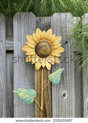 A large Sunflower made out of metal,complete with green leaves. The flower is mounted to the side of a light brown backyard fence.