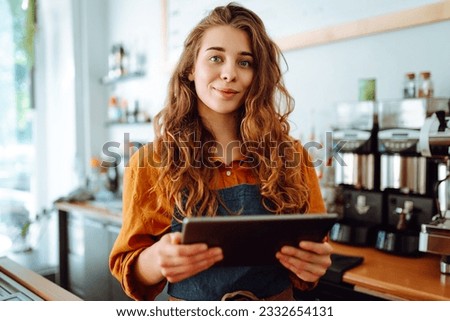 Beautiful woman owner stands behind the counter of a coffee shop. A barista with a digital tablet takes an order. Business concept. Takeaway food. Royalty-Free Stock Photo #2332654131