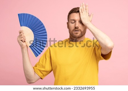 Sweaty tired middle aged man touching forehead using paper fan suffer from heat, feels sluggish. Displeased guy cooling in hot summer weather, isolated on studio pink background. Overheating concept Royalty-Free Stock Photo #2332693955
