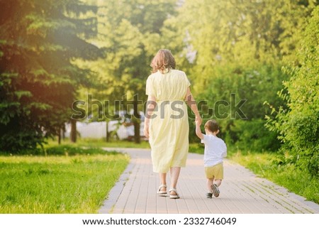 A child takes steps holding a mother woman by the hand along a path in nature. Happy baby with mom on a walk in the summer park. Kid aged about two years (one year eleven months) Royalty-Free Stock Photo #2332746049