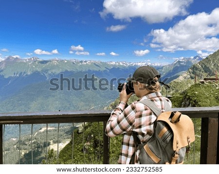 young stylish photographer woman taking photo of mountains standing at scenic viewpoint at Rosa peak, Sochi, Russia