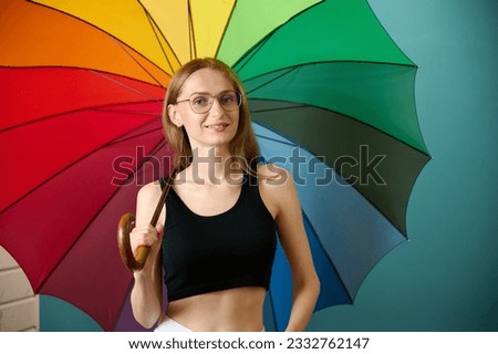Happy caucasian female in black top and white pants under rainbow colored umbrella posing at studio background, free copy space