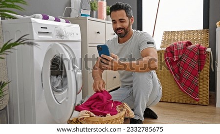Young hispanic man using smartphone washing clothes smiling at laundry room