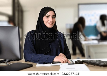 Emirati at school classroom with students or co-teacher and write board at background. Arab woman sitting at a table with desktop screen monitor