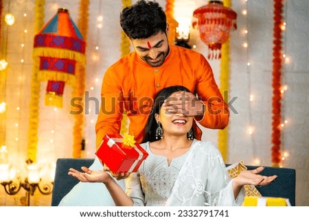 Happy indian man by closing eyes giving surprise gift to wife during festival celebration at home - concept of relationship bonding, togetherness and raksha Bandhan. Royalty-Free Stock Photo #2332791741