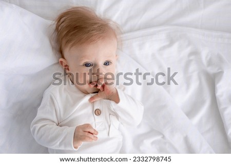 smiling baby 6 months old blond boy lies on a white bed in a bright bedroom and licks his finger in a cotton bodysuit, the concept of children's goods