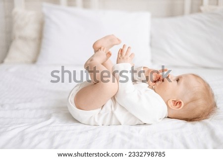 smiling baby 6 months old blond boy lies on a large bed in a bright bedroom and plays with his legs in a cotton bodysuit, the concept of children's goods