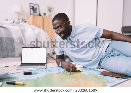 Positive African American man in casual clothes reading map while leaning on floor near laptop with blank screen at home