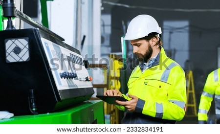Caucasian male worker of plant in helmet and yellow uniform standing at controlling point with tablet device in hand and running michine. Indoors. Handsome man in hardhat at controller in production. Royalty-Free Stock Photo #2332834191