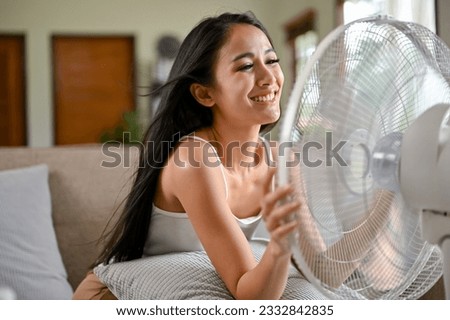 A happy and pretty Asian woman breathes fresh air from an electric fan while resting on a couch in her living room on a hot summer day. Royalty-Free Stock Photo #2332842835