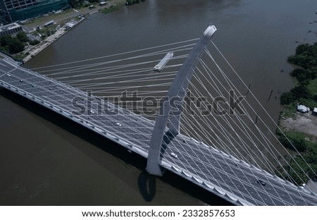 High aerial angle of suspension bridge over river with road traffic in dramatic light. Cables of bridge create strong graphic lines. A boat passes through picture.