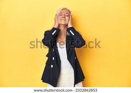 Middle-aged Caucasian woman in business jacket, yellow studio laughs joyfully keeping hands on head. Happiness concept.
