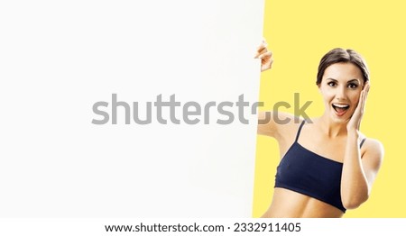 Photo of excited beautiful brunette woman in fitness wear showing blank banner paper signboard with copy space area. Fit girl with happy wide open mouth, peek out mock up white placard, over yellow.