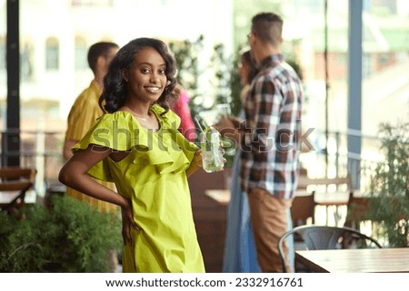 Beautiful, young african woman in stylish clothes meeting with friends at cafe, having funny, joyful time. Blurred people on background. Concept of friendship, party, communication, alcohol drinks