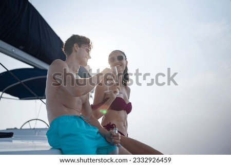 Low angle view, selective focus of an attractive Caucasian couple in short pants and bikini, wearing sunglasses, smiling clinking wine glasses together on a yacht with a bright white sky in background