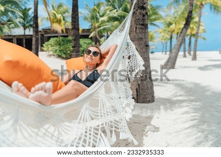 Smiling middle-aged woman relaxing lying in wicker hammock in palm trees grove on white sandy beach on Mauritius coast and enjoying . Exotic countries vacation and mental health concept image Royalty-Free Stock Photo #2332935333