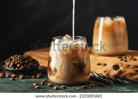 Ice coffee in a tall glass with cream poured over, ice cubes and beans on a dark concrete table. Cold summer drink with tubes on a black background with copy space. Royalty-Free Stock Photo #2333005681