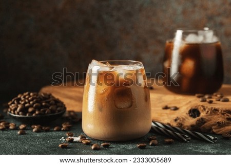 Ice coffee in a tall glass with cream poured over, ice cubes and beans on a dark concrete table. Cold summer drink with tubes on a black background with copy space. Royalty-Free Stock Photo #2333005689