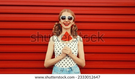 Summer portrait of happy smiling young woman with fresh juicy fruits, lollipop or ice cream shaped slice of watermelon wearing heart shaped sunglasses with cool girly hairstyle on red background