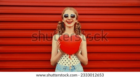 Portrait of happy smiling cute girl with big red heart shaped balloon and girly hairstyle having fun wearing sunglasses on background