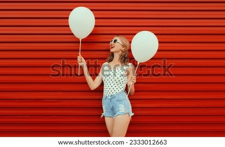 Happy cheerful young woman having fun with balloon wearing white heart shaped sunglasses, shorts on red background