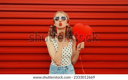 Portrait of happy cute girl with big red heart shaped balloon and girly hairstyle blows her lips sends air kiss wearing sunglasses on background