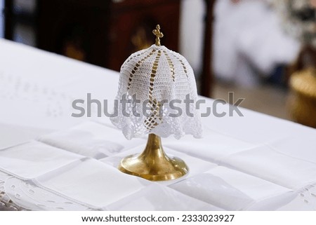 golden metal chalice or goblet with symbol of the cross of Christ, covered with white embroidered fabric on the altar - ampulla Royalty-Free Stock Photo #2333023927