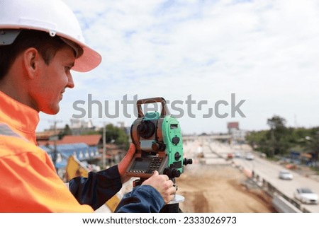 Site young man engineer operating his instrument during roadworks looking at camera. Builder using total positioning station tachymeter on construction site for new road setting out