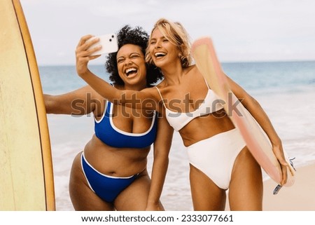 Summer surf fun comes to life as female surfers capture a selfie on the beach, with shared excitement and the spirit of summer filling the air. Two women in bikinis having fun on a surfing holiday.