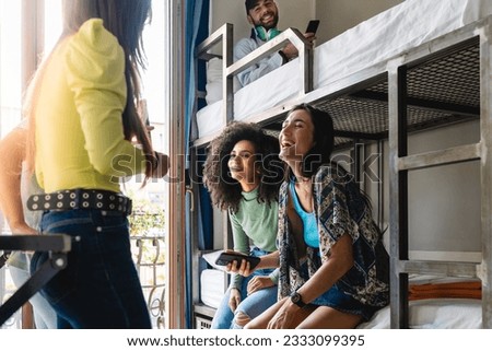 Two women, Caucasian and Brazilian, laughing with a smartphone, surrounded by friends in a hostel room, boy on upper bunk laughs along Royalty-Free Stock Photo #2333099395