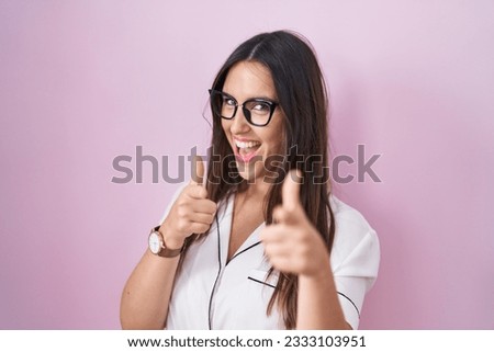 Young brunette woman wearing glasses standing over pink background pointing fingers to camera with happy and funny face. good energy and vibes. 