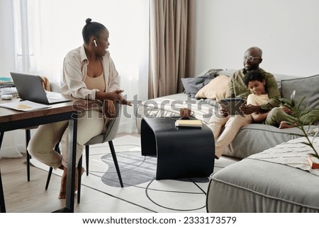 African American mother working online on laptop at home while her family having fun with tablet pc on sofa in background