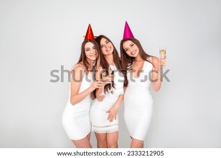 Three young woman in elegant dresses having fun, smiling, dancing and drinking champagne in studio on white background. Christmas party celebration concept.