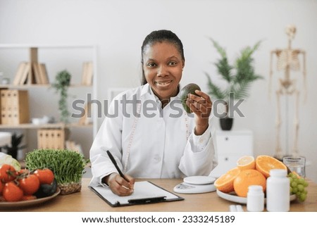 Portrait of smiling multicultural lady wearing white coat with avocado in hand posing at writing desk. Cheerful expert in food and nutrition selecting food for special diet in doctor's workplace.