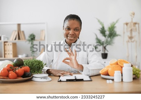 Portrait of attractive adult woman in doctor's coat posing at office desk with healthy food and medicine on it. Multicultural dietitian planning and conducting nutritional programs showing sign ok