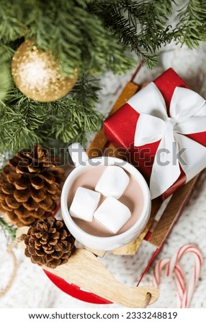 close-up of christmas time themed decorations by the tree- toy sled with hot chocolate filled cup and marshmallows, nicely wrapped present, candy canes and tree cones, cozy knitted throw in the backgr
