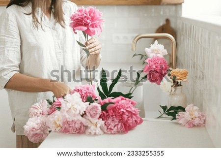 Woman arranging beautiful peonies in vase at sink with brass faucet in new modern kitchen. Female decorating flowers on background of granite countertop, hands with flowers close up